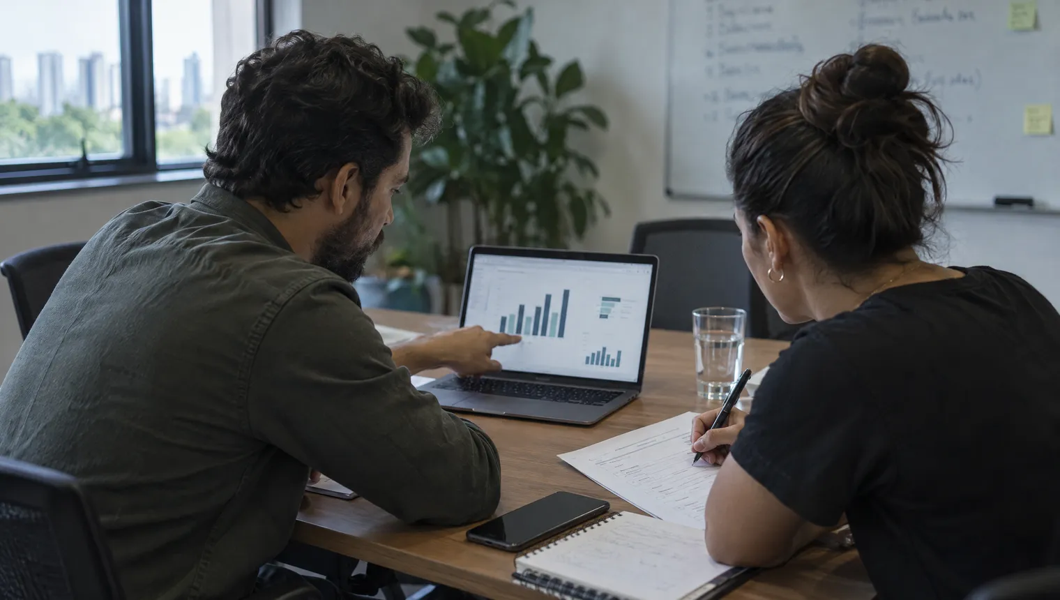 Numa sala de reunião, um homem aponta para um gráfico de barras no notebook enquanto uma mulher marca itens numa lista impressa.