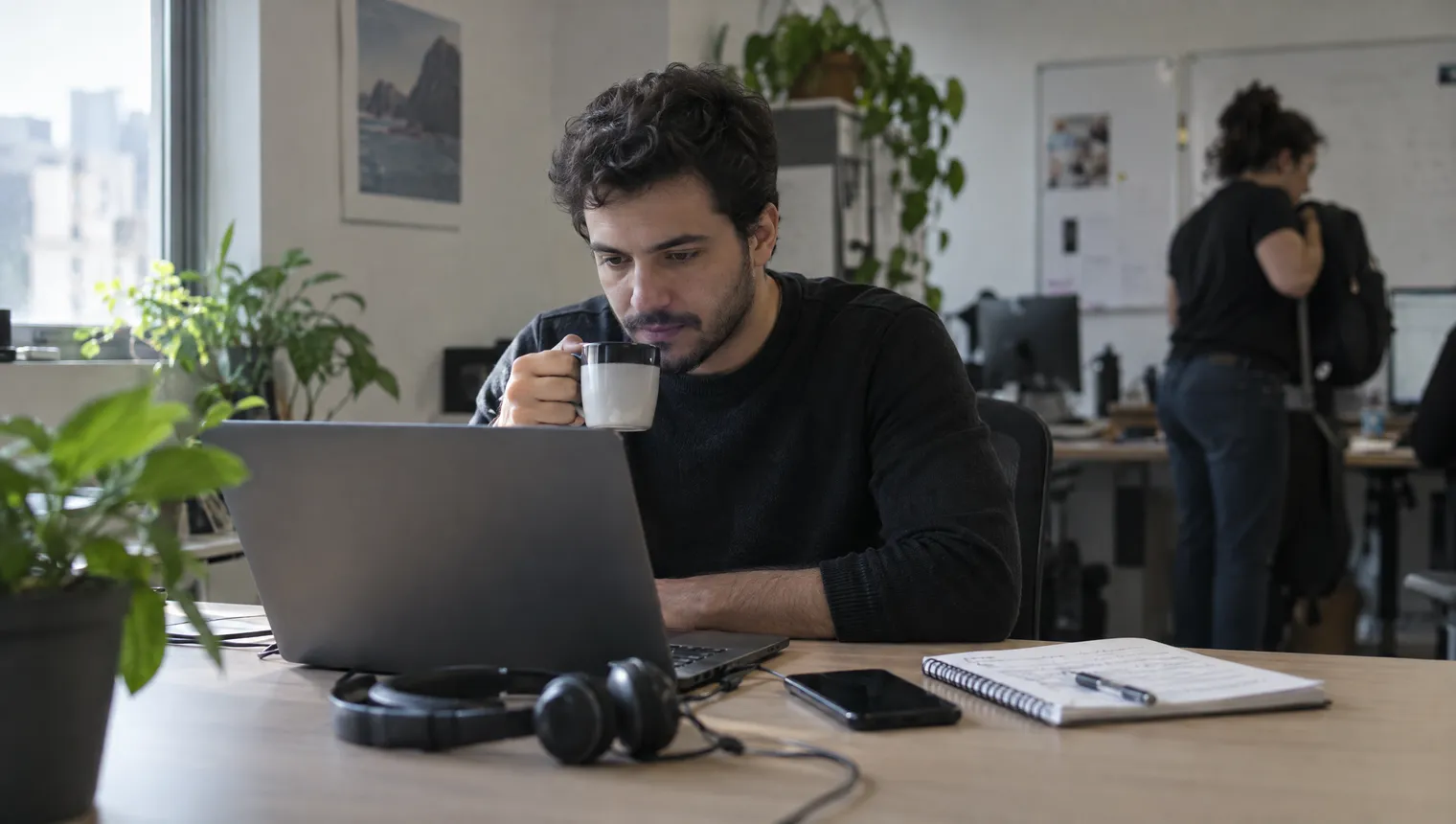 De manhã, numa sala de atendimento, um atendente com uma xícara de café lê no notebook as conversas que chegaram durante a noite; um fone de ouvido e o celular estão sobre a mesa.